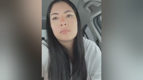 Media: Video of a young woman with long, straight black hair, wearing a white top, sitting in a car. She has light skin and is looking directly at the camera with a neutral expression.