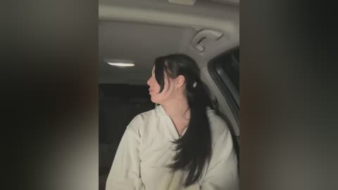 Media: Video of a woman with long, dark hair in a ponytail, wearing a white robe, standing inside a dimly lit car.