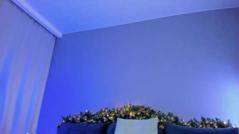 Media: A video of a cozy living room corner with a gray wall, a blue ceiling, and a festive garland of greenery and golden lights across a dark sofa.