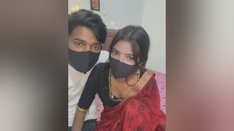 Media: A video of a young South Asian couple in a cozy room. The man, with short black hair, wears a white T-shirt and black mask, while the woman, with dark hair in a bun, sports a red sari with a black mask, gold earrings, and a necklace.