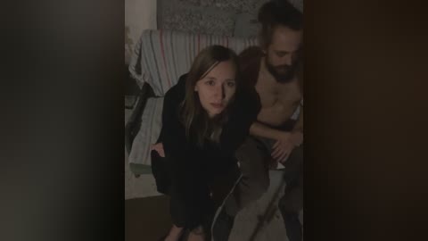 Media: Video of a dark-haired woman in a black robe, crouching near a shirtless man with long hair, in a dimly lit bedroom with a bed and striped blankets.