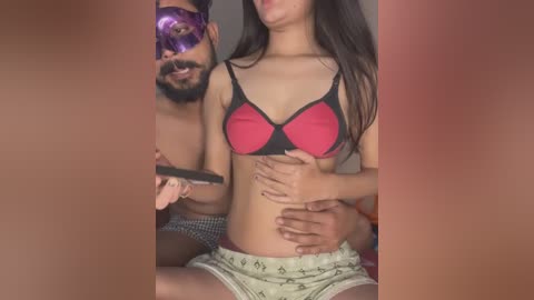 Media: Video of a man in a purple mask, holding a smartphone, and a woman in a black and red bra, white shorts, and long dark hair, sitting on a bed.