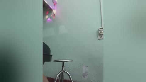 A video of a bathroom with a pastel green wall, a metal stool, and a dangling string of colorful lights. The background features a white electrical outlet and a faintly visible sink. The image has a soft, diffused lighting effect.