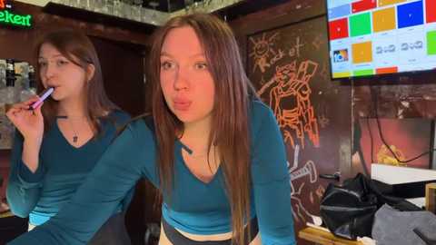 Media: Video of two young women with long brown hair, wearing teal tops, in a lively bar, one licking a lollipop, another puckering her lips. Background features colorful chalkboard art, bar stools, and a TV screen.