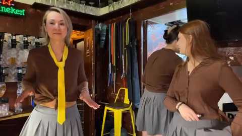 Media: Video of a stylish woman in a brown shirt with a yellow tie, gray skirt, and a tattooed arm, standing in a dimly lit bar with neon signs, wooden walls, and a yellow stool.