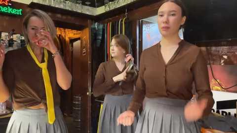 Media: Video of a woman with glasses, brown hair, in a brown top, gray skirt, and yellow tie, dancing in a cozy bar with another woman, reflecting in a mirror.