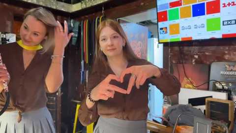 Media: Video of two young women in a bar, one with blonde hair and a yellow necklace, the other with brown hair, both making heart shapes with their hands. Background features colorful lights, shelves, and a \"Magnum\" sign.