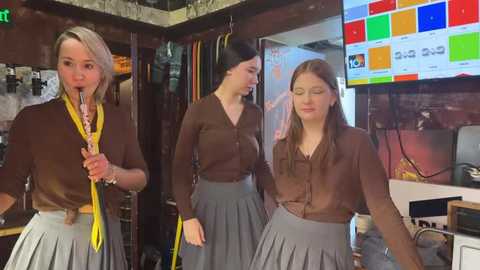 Media: Video of three women in brown blouses and pleated grey skirts, dancing and smiling, in a modern office with a colorful digital screen in the background.