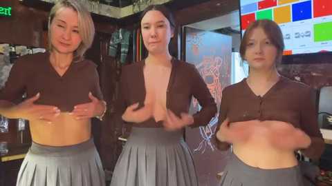 Media: Video of three women in a bar, topless, wearing low-cut, brown cardigans and gray pleated skirts, holding their breasts. Background shows a dimly lit interior with bar paraphernalia.