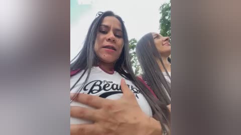 Media: Video of a woman with long, straight black hair wearing a white \"Bear\" t-shirt, touching her chest, standing outside with blurred background of trees.