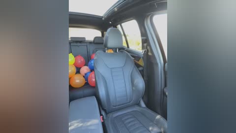 Media: Video of a car interior with black leather seats, featuring a pile of colorful balloons (red, blue, yellow) on the backseat, illuminated by daylight.