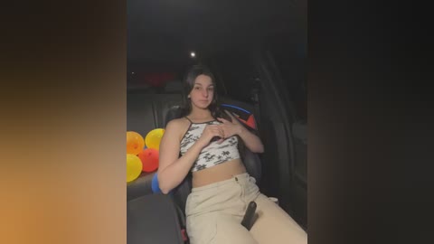 Media: Video of a young woman with long dark hair, wearing a white crop top and beige pants, sitting in a car at night, surrounded by colorful balloons.