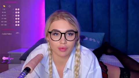 Media: Video of a young blonde woman with braided hair, wearing black-rimmed glasses and a white blouse, sitting on a bed with blue curtains, holding a microphone, in a dimly lit room.