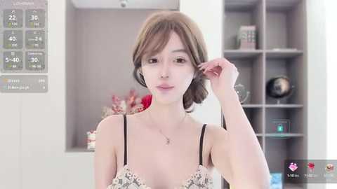 Media: Video of a young woman with light skin, short brown hair, wearing a white lace bra, adjusting her hair in a modern, well-lit living room with white walls and a gray bookshelf.