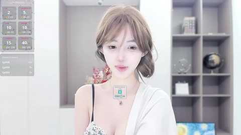 Media: A video of a young, slender East Asian woman with fair skin and shoulder-length brown hair, wearing a white robe and a floral-patterned bra, standing in a modern, minimalist room with gray shelves and a clock.