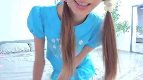 Media: Video of a smiling, light-skinned girl with long brown pigtails in a blue dress, leaning on a bed in a bright, minimalistic bedroom.