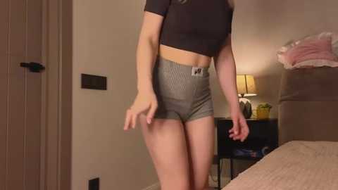 Media: Video of a slender, light-skinned woman in black crop top, high-waisted gray plaid shorts, standing in a dimly-lit bedroom with beige walls, bed, lamp, and closed door.