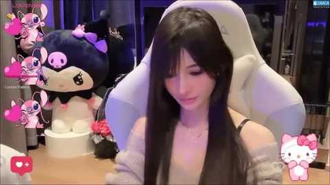 Video of a young woman with long black hair, wearing a gray sweater, seated at a desk, surrounded by plush toys and a Hello Kitty figure, in a cozy, dimly lit room.