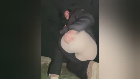 Media: Video of a person in a black jacket and pants, lying on a grassy ground, with a large, round, red and white bruise on their stomach. The background is dark, suggesting nighttime or dim lighting.