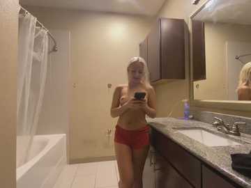 Media: Video of a blonde woman, topless with red shorts, standing in a bathroom with beige walls, granite countertop, and mirror.