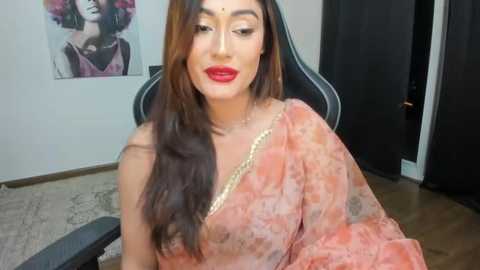 Media: Video of a South Asian woman with long, wavy brown hair, wearing a pink floral saree with gold embroidery, sitting in a gaming chair, in a brightly lit room with a floral print wall and a closed door.