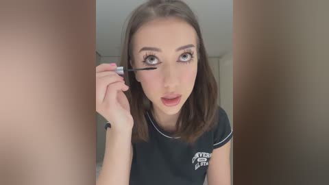 Media: A video of a young woman with fair skin, brown hair, and light makeup, applying eyeliner in a bathroom with beige walls. She wears a black T-shirt.