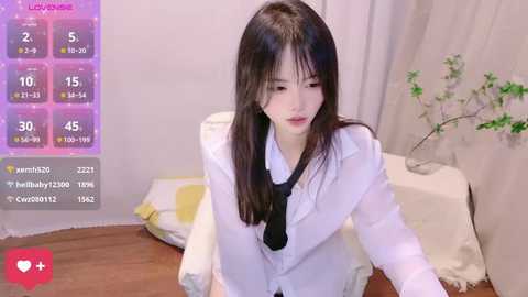 Media: Video of an East Asian woman with long black hair, wearing a white lab coat and black tie, leaning over a wooden table in a minimalist room with a plant.