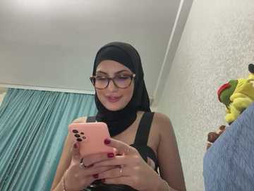 Media: Video of a fair-skinned woman with glasses, black hijab, and black tank top, taking a selfie with a pink iPhone. Background features a white patterned wall, teal curtains, and a stuffed toy.