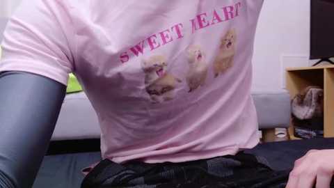 Media: A video of a person wearing a pink T-shirt with \"SWEET HEART\" in pink letters, featuring three cartoon dogs, and gray compression sleeves, sitting on a couch. The background includes a TV stand and a green bag.