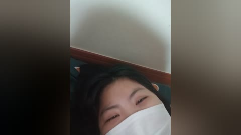 Media: Video of an Asian woman with short black hair and fair skin, wearing a white surgical mask, lying on a dark green bed, with a wooden headboard in the background.