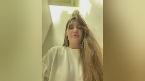 Media: Video of a young woman with long, straight blonde hair, wearing a white shirt, standing in a dimly lit hallway with cream-colored walls. Her eyes are closed, and she appears relaxed.