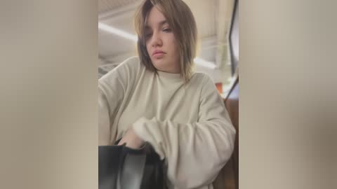 Media: A video of a young woman with shoulder-length brown hair, wearing a cream-colored sweater, sitting indoors, holding a black bag, with blurred background featuring a ceiling and wooden furniture.