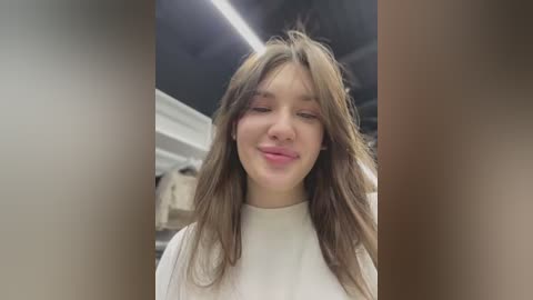 Media: A video of a young woman with long, wavy brown hair, wearing a white t-shirt, smiling and sticking her tongue out, taken indoors under bright, overhead lights.