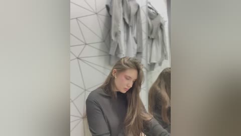 Media: A video of a young woman with long, straight brown hair, wearing a black turtleneck, standing in a modern bathroom with gray geometric tiles and hanging gray robes.