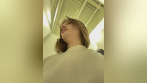 Media: Video of a young woman with light skin and shoulder-length brown hair, wearing a white shirt, looking upward in a dimly lit, industrial space with white ceiling beams.