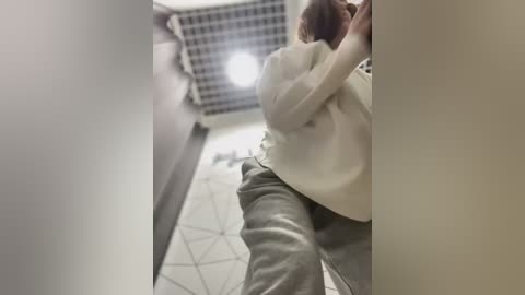 Media: A video of a person, likely a woman, with light skin, red hair, and wearing a beige sweater, crouching in a narrow, tiled hallway with a checkered ceiling light.