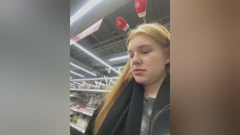 Media: Video of a young blonde woman with fair skin in a supermarket, wearing a black jacket, eyes closed, with red gloves dangling above her head.