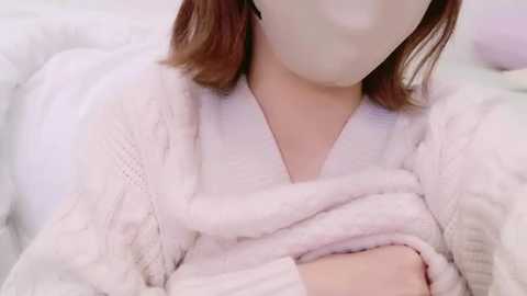 Media: Video of a person wearing a fluffy white sweater, with light brown hair, blurred face, sitting on a white couch, creating a cozy, soft, and serene atmosphere.