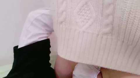 Media: A close-up video of a person wearing a cream cable-knit sweater over a white shirt, with a black jacket sleeve partially visible. The background is blurred, focusing on the texture of the sweater.