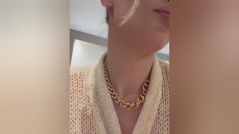 Media: Video of a close-up of a woman's neck and chin, wearing a gold chain necklace and a beige, knitted sweater. The background is blurred, featuring a neutral-toned wall.