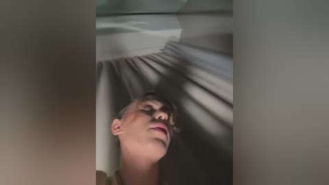 Media: A video shows a young woman with short, light brown hair and fair skin, lying on her back in a dimly lit, modern room. She has a serene expression, looking upwards, with sunlight streaming through blinds creating shadows on the ceiling.