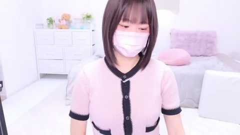 Media: A video of an East Asian woman with shoulder-length black hair, wearing a sheer pink top, white face mask, and seated in a white room with light furniture.