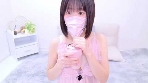 Media: Video of a young Asian woman with short black hair, wearing a pink face mask and revealing lingerie, sitting on a gray carpeted floor in a minimalist white room.