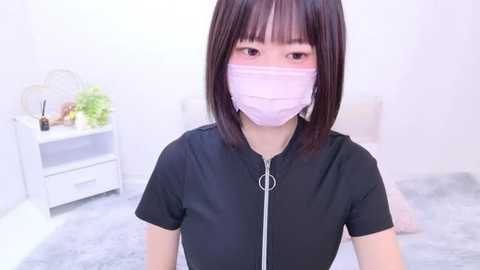 Media: Video of an East Asian woman with straight black hair, wearing a pink surgical mask and black short-sleeve top, standing in a minimalist white room with a white nightstand and plants.