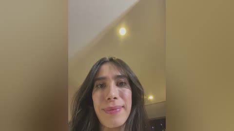 A video of a smiling young woman with long, dark hair, light skin, and a slight tan. She is indoors, with beige walls and recessed lights in the background.