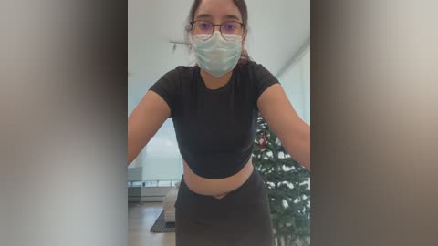 Media: Video of a young woman with light skin and glasses, wearing a black crop top and leggings, a face mask, and standing in a modern room with a Christmas tree in the background.