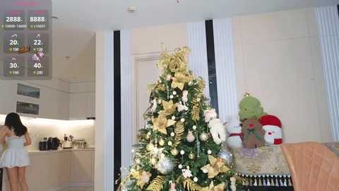 Media: Video of a modern living room with a decorated Christmas tree, plush toys, and a person in a white dress near a kitchen counter.