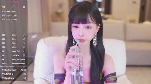 Media: A video of an Asian woman with long black hair and bangs, wearing a strapless dress, drinking from a glass bottle in a modern living room.