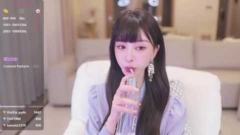 Media: Video of an East Asian woman with long black hair and fair skin, wearing a light blue dress, drinking water in a modern, indoor setting with a blurred background.