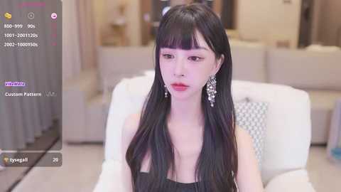 Media: A video of a young East Asian woman with long black hair and red lipstick, wearing a black dress and silver earrings, seated indoors on a white couch.